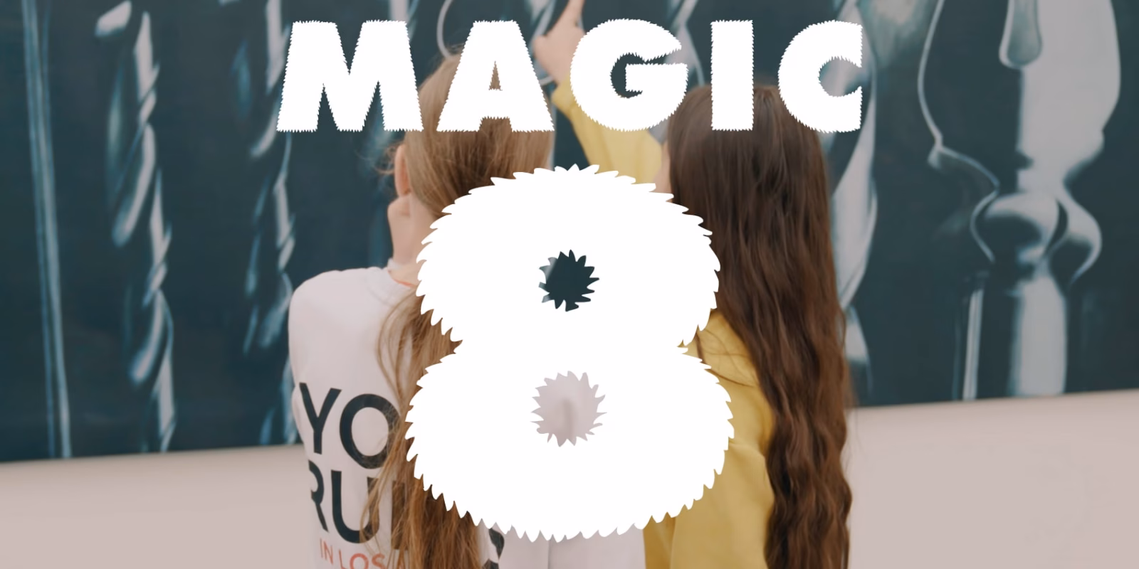 Twee meisjes met lang haar staan met hun rug naar de camera dicht bij een grote muurschildering en steken hun armen omhoog terwijl ze naar het kunstwerk wijzen. Op de voorgrond staat in grote witte letters “Magic 8”.