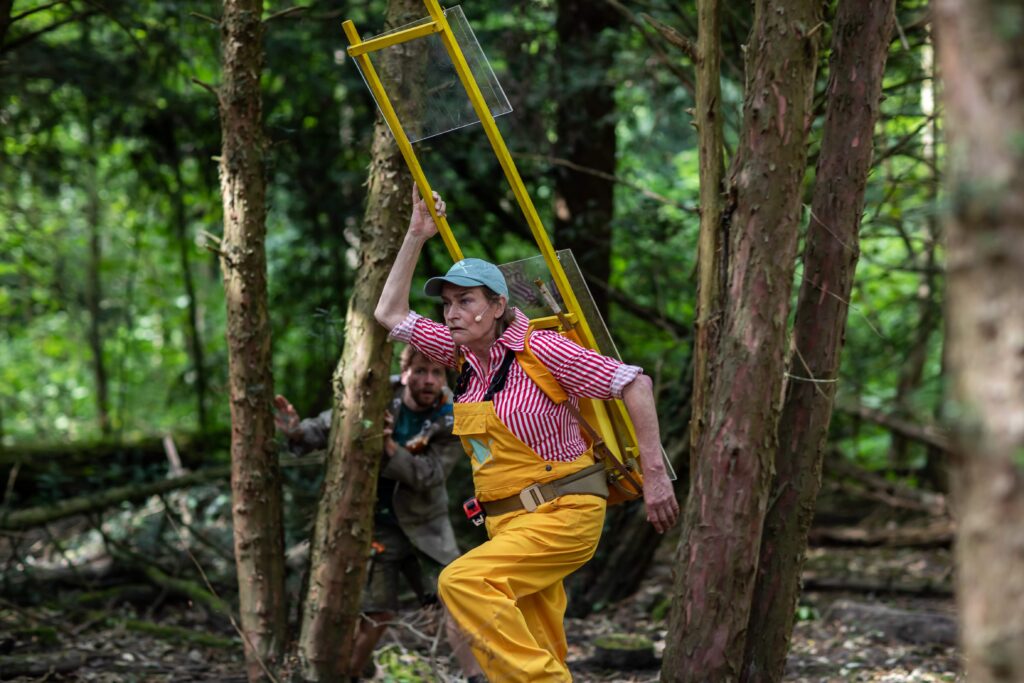 Speler in rood-wit overhemd en gele tuinbroek loopt met een ladder door het bos terwijl een andere speler haar vanuit de bomen observeert.