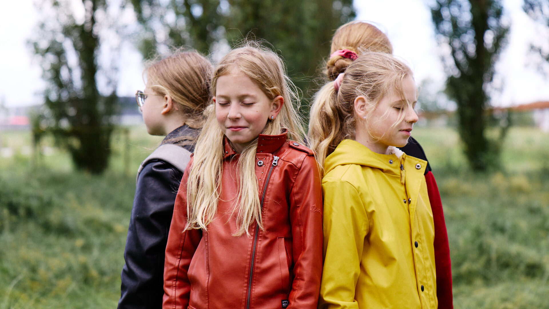 Vier kinderen staan buiten rug-aan-rug in een kleine kring. De kinderen hebben hun ogen dicht. Op de voorgrond staan twee meisjes: één in een rood leren jasje en één in een gele regenjas. Bomen en een groen veld vormen de achtergrond.